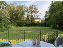 Ein Garten mit großer, gepflegter Rasenfläche, umgeben von hohen Bäumen und Sträuchern. Im Vordergrund eine Terrasse mit dekorativem Metallgeländer, einem Tisch mit Tischdecke und zwei Kerzengläsern. Im Hintergrund ein Kinderspielplatz mit bunter Rutsche.