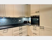 Moderne Küche mit cremefarbenen Einbauschränken, schwarzer Arbeitsplatte und einer Rückwand aus glänzenden, grünlich-blauen Fliesen im U-Bahn-Stil. Integriert sind ein Backofen und ein Kochfeld, mit kleinen Elektrogeräten auf der Arbeitsplatte.