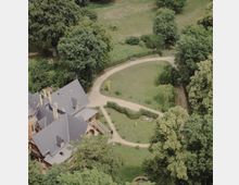 Vogelperspektive auf ein historisches Herrenhaus mit grauem Schieferdach, umgeben von einem weitläufigen Garten mit Rasenflächen, geschwungenen Wegen, Hecken und großen Bäumen. Im Garten befinden sich Bänke und eine kleine Statue.