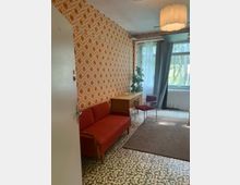 Kleines Wohnzimmer mit orange gemusterter Tapete, rotem Sofa, kleinem Holztisch und Stuhl sowie gemustertem Fliesenboden mit Teppich. Am Fenster mit Vorhängen steht eine Pflanze; an der Decke hängt eine große, runde Deckenlampe.