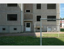 Wohnblock mit grauer, rauer Fassade und mehreren offenen Fensteröffnungen, die auf einen verlassenen oder unvollständigen Zustand hindeuten. Im Vordergrund Rasen, ein Wäschegerüst und ein gepflasterter Weg; rechts sind Briefkästen und ein kleines Geländer sichtbar.