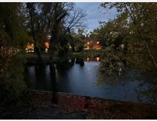 Abendliche Ansicht eines kleinen Teichs oder Flussarms, umgeben von hohen Bäumen mit herbstlichem Laub. Im Hintergrund sind beleuchtete Gebäude und Straßenlampen zu sehen, deren Licht sich auf der dunklen Wasseroberfläche spiegelt.