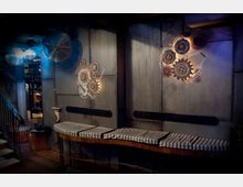 Industriell gestalteter Innenraum mit grauen Metallwänden, rustikalen Zahnrädern als Dekoration und Holzbänken mit gestreiften Polstern. Im Hintergrund sind Treppen und metallische Hängelampen sichtbar.