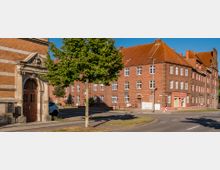 Straßenecke mit Backsteingebäuden und roten Dächern im Stil traditioneller Wohnarchitektur. Im Vordergrund ein Baum, eine breite Straße, Gehwege und ein verzierter Eingang mit steinernem Türrahmen.