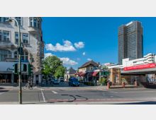 Straßenkreuzung in einer Stadt mit historischen Gebäuden auf der linken Seite und flachen Geschäftsgebäuden, darunter ein Restaurant, auf der rechten Seite. Im Hintergrund ragt ein modernes Hochhaus in den blauen Himmel, und Bäume säumen einen kleinen Straßenabschnitt.