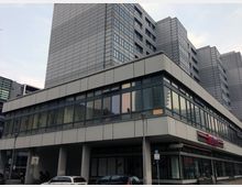 Mehrstöckiges modernes Gebäudekomplex aus Beton und Glas mit großen Fenstern und kantigem Design. Im Erdgeschoss befinden sich Geschäfte, darunter ein Laden mit rotem Schriftzug, und davor parkende Autos.