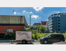 Straßenansicht mit einem älteren, grauen Gebäude links und einem mehrstöckigen Wohnhaus rechts; dazwischen befinden sich ein bewachsener Zaun und ein leeres Grundstück. Im Vordergrund stehen ein Anhänger mit Graffiti und ein schwarzer PKW, daneben ein Straßenschild mit der Aufschrift „Selerweg“.