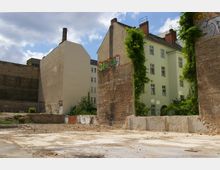 Freifläche in einer Stadt mit sichtbaren Überresten eines abgerissenen Gebäudes, darunter Mauerfragmente mit Graffiti. Im Hintergrund stehen hell verputzte Wohnhäuser mit mehreren Fenstern, teilweise von Kletterpflanzen bewachsen.