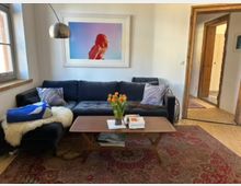 Gemütliches Wohnzimmer mit dunkler Eckcouch, Kissen und Decken sowie einem niedrigen Holzcouchtisch mit Tulpen und Büchern auf einem gemusterten Teppich. An der weißen Wand hängt ein großes gerahmtes Bild; links fällt Tageslicht durch ein Fenster, rechts führt ein rustikaler Holztürrahmen in einen Flur mit Holzdielen.