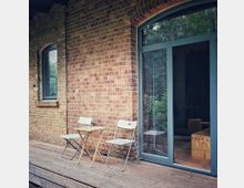 Backsteinfassade mit einer großen Glastür und einem schmalen Fenster mit blauen Rahmen. Davor stehen zwei Klappstühle und ein kleiner Holztisch auf einer Holzterrasse, umgeben von einer natürlichen Umgebung.
