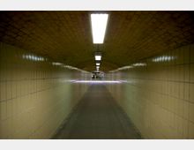 Langer, gewölbter Fußgängertunnel mit gelb gefliesten Wänden und heller Deckenbeleuchtung, die in gleichmäßigen Abständen angebracht ist. Am Ende des Tunnels ist ein offener Bereich mit weiterer Beleuchtung und Personen sichtbar.