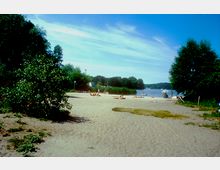 Sandstrand mit einzelnen Menschen, die sonnenbaden, umgeben von Bäumen und Büschen; im Hintergrund ein See mit Schilf am Ufer sowie vereinzelten Booten und Segeln auf dem Wasser.