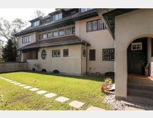 Großes Wohnhaus im traditionellen Stil mit heller Putzfassade und roten Ziegeln auf dem Dach. Im Vordergrund ein gepflegter Garten mit Rasenfläche, gepflastertem Weg und einer Veranda mit Rundbogen.