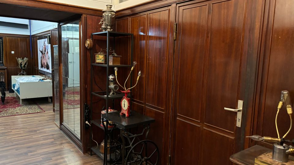 Innenraum mit dunkler Holzvertäfelung und Holzboden, daneben eine Glastrennwand und eine Tür. Im Vordergrund stehen Regal und Tische mit alten technischen Geräten und Messinstrumenten, im Hintergrund ist ein weiterer Raum mit Teppich und Möbeln zu sehen.