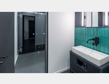 Modernes Badezimmer mit einer weißen Waschschale auf einem dunklen Waschtisch, einer Wandverkleidung aus türkisfarbenen Fliesen und integrierter Beleuchtung über dem Spiegel. Im Hintergrund sind gläserne Duschabtrennungen und ein dunkler Boden zu sehen.