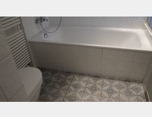 Badezimmer mit einer weißen Badewanne, gefliesten Wänden in Weiß, einem dekorativ gemusterten Fliesenboden und einem Heizkörper aus weißen Rohren neben einer Toilette.