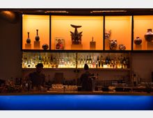 Eine Bar mit einer beleuchteten blauen Theke; darüber Regale mit einer Vielzahl von Spirituosenflaschen und leuchtend orangefarbenen Dekorationselementen, darunter Vasen, Masken und eine Ikone. Zwei Personen arbeiten hinter der Theke, die mit Gläsern und Zutaten ausgestattet ist.