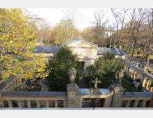 Historischer Friedhof mit einer aufwendig gestalteten Familiengruft, umgeben von Bäumen und Herbstlaub. Die Gruft ist mit Statuen von Engeln, einem Kreuz und grünen Sträuchern geschmückt, während eine Balustrade den Bereich umrahmt.