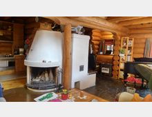 Gemütliches Wohnzimmer in einem Blockhaus mit sichtbaren Holzstämmen und flachen Holzdeckenbalken. Im Vordergrund ein weißer Kachelofen mit Kaminöffnung, daneben eine offene Küche und im Hintergrund ein Arbeitsbereich mit Holzschreibtisch und Regalen.