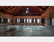 Ein leerer, heller Raum mit gewölbten Fenstern und offenen Holzläden, der durch eine sichtbare Holzbalkendecke und unverputzte Ziegelwände geprägt ist. Der Boden ist teilweise mit grobem Material belegt, und das Licht fällt durch die Fenster und erzeugt markante Schatten.