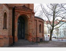 Backsteinbau mit rundbogigen Fenstern und einem Eingangsportal mit einem Rundbogen, das über eine kleine Treppe erreichbar ist. Im Hintergrund Häuserfassaden und kahle Bäume in städtischer Umgebung.