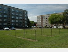 Mehrstöckige Wohnblöcke aus Beton mit zahlreichen Fenstern, die von einer Grünfläche umgeben sind. Im Vordergrund stehen Metallgestelle, möglicherweise für Wäscheleinen, während auf der Wiese parkende Autos und ein Baum zu sehen sind.