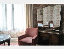 Ein Zimmer im Retro-Stil mit floral gemusterter Tapete, einem roten Polstersessel, einem Holztisch mit Glasplatte und einer Kommode mit dreiteiligem Spiegel. Im Hintergrund sind lichtdurchlässige Vorhänge und ein Teil eines Bettes sichtbar.