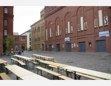 Freier Platz mit Kopfsteinpflaster vor dem Gebäude eines alten Industriekomplexes aus rotem Backstein mit der Beschriftung „Kesselhaus“. Im Vordergrund stehen aufgestellte Bierbänke, links parken Fahrräder, und im Hintergrund sind weitere Gebäude zu sehen.