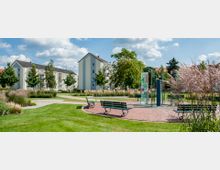 Ein gepflegter Park mit Grünflächen, Bäumen, Ziergräsern und gepflasterten Wegen, umgeben von mehrstöckigen Wohngebäuden. Im Vordergrund sind Sitzbänke und ein Kunstwerk aus gläsernen, bunten Paneelen sichtbar.