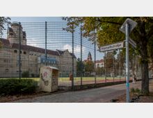 Straßenszene mit einem eingezäunten Sportplatz im Vordergrund, umgeben von Bäumen und Gehwegen, sowie einem historischen Gebäude mit roten Dächern und einer Turmstruktur im Hintergrund; ein Straßenschild mit der Aufschrift "Homuthstraße" ist rechts im Bild sichtbar.