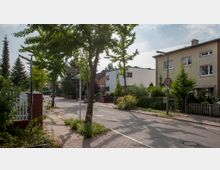 Ruhige Wohnstraße mit Gehweg, gesäumt von Bäumen und gepflegten Vorgärten. Links und rechts stehen Ein- und Mehrfamilienhäuser, ein Straßenschild mit der Aufschrift „Almweg“ und ein Verkehrsschild mit einer Geschwindigkeitsbegrenzung von 30 km/h sind sichtbar.