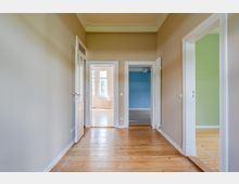 Heller Flur in einer Wohnung mit hohen Decken, Stuckleisten und Holzdielenboden. Mehrere weiße Türrahmen führen in angrenzende Räume, darunter ein blau gestrichener Raum und ein Raum mit grüner Wand sowie ein Fensterbereich am Ende des Flurs.