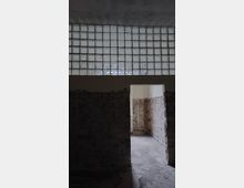 Ein leerer und unrenovierter Raum mit freigelegten Ziegelwänden und einem Betonboden. Oberhalb der Türöffnung befindet sich eine große Fläche aus Glasbausteinen, die gedämpftes Licht hereinlässt.