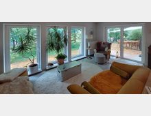 Ein modernes Wohnzimmer mit großen Glasfenstern, die den Blick auf einen Garten mit Bäumen ermöglichen. Im Raum befinden sich ein gelbes Sofa, ein Ohrensessel, Zimmerpflanzen in Töpfen und ein Couchtisch auf einem runden Teppich, hinter der Fensterfront ist eine Terrasse mit Holztisch und Stühlen sichtbar.