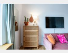 Wohnzimmer-Ecke mit einem rattanartigen Schrank, auf dem zwei Kakteen in Terrakotta-Töpfen und eine Tischlampe stehen. Daneben befindet sich ein rosafarbenes Sofa mit farbigen Kissen, eine Wand mit einem Flachbildfernseher sowie ein Spiegel und ein türkisfarbener Vorhang.