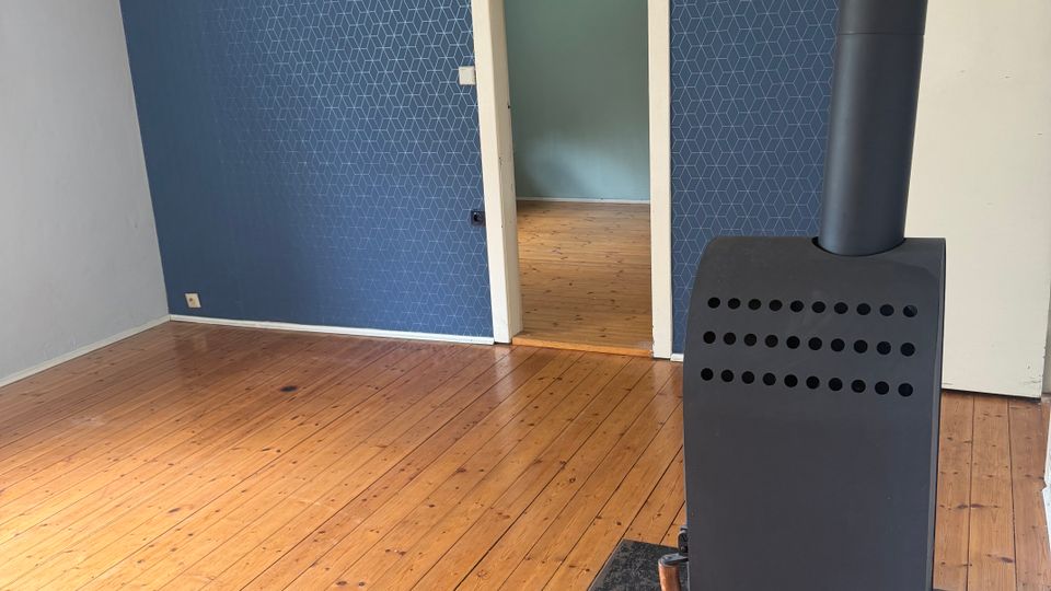 Leerer Wohnraum mit breiten Holzdielenboden und einer blauen, geometrisch gemusterten Akzentwand. Rechts steht ein schwarzer Kaminofen mit Ofenrohr; in der Mitte führt eine Türöffnung in einen angrenzenden Raum.