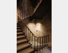 Eine Betonwendeltreppe mit schwarzem Metallgeländer in einem schlichten, rauen Innenraum. Die Wände sind unverputzt, und eine kleine, metallene Tür befindet sich unten an der Treppe.