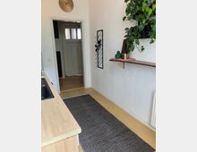 Schmale Küche mit hellen Wänden und gelbem Bodenbelag, an einer Seite eine Holz-Arbeitsplatte mit Spüle, gegenüber eine Wand mit Regal, Weinregal und hängenden Pflanzen. Im Hintergrund eine Tür mit Glaseinsätzen, die zu einem angrenzenden Raum führt.