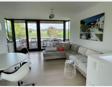 Modernes Wohnzimmer mit großer Glasfront, die auf eine Terrasse und eine grüne Landschaft blickt. Im Raum befinden sich ein Ecksofa, ein runder Esstisch mit Stuhl sowie minimalistische Dekorationen; an der Wand hängt ein großes gerahmtes Bild.