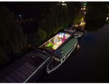 Nachtansicht eines Bootsanlegers mit beleuchteten Hausbooten an einem Fluss, umgeben von Bäumen. Auf einem der Boote befindet sich eine bunte Lichtinstallation mit lebhaften Farben.