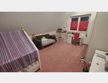 Ein Kinderzimmer mit Dachschräge, rotem Teppichboden und weißer Wandgestaltung. Das Zimmer enthält ein Schlafbett mit lila-gestreiftem Stoffdach, ein graues Sofa, ein Schreibtisch mit rotem Stuhl vor einem doppelflügeligen Fenster mit roten Plissees sowie Spielzeug, Regale und Dekorationen an den Wänden.