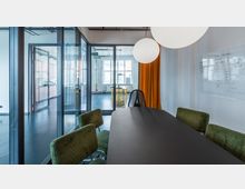 Modernes Besprechungszimmer mit großem dunklem Tisch, grünen Polsterstühlen und gläsernen Trennwänden; ein orangefarbener Vorhang und eine beschriftete Weißwandtafel zieren den Raum. Im Hintergrund sind angrenzende Räume durch Glaswände sichtbar, darunter eine Küche und ein Bereich mit Fenstern, die viel Tageslicht hereinlassen.