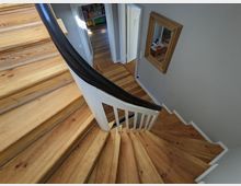 Blick von oben auf ein geschwungenes Holztreppenhaus mit hellen Stufen, weißem Geländer und dunklem Handlauf. Unten führt ein Flur mit Holzboden zu mehreren Türen; an der Wand hängt ein rechteckiger Spiegel mit Holzrahmen.