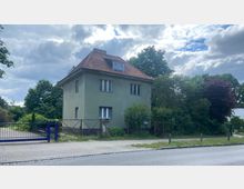 Zweistöckiges Wohnhaus mit grauer Fassade und rotem Ziegeldach, umgeben von dichtem Grün. Vor dem Haus befinden sich ein gepflasterter Gehweg, ein blaues Tor und Gitterzäune.