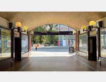 Arkadenpassage mit gelbem Kachelgewölbe und beidseitigen Schaufenstern. Im Hintergrund sind gepflasterte Wege, Bäume und ein modernes Gebäude mit Glasfassade zu sehen, sowie ein Wegweiser mit mehreren Ausgängen.