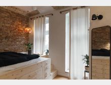 Schlafzimmer mit einem Podestbett aus hell gemasertem Holz, eingebauten Schubladen und einer Stufenaufgang. Links eine Wand aus Ziegelsteinen, rechts zwei hohe Fenster mit halbtransparenten weißen Vorhängen, daneben ein stehender Spiegel.