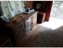 Ein rustikaler Innenraum mit einem Holzboden, einer hölzernen Kommode mit Teekanne, Tassen und einer Metallkörbchen-Dekoration. Im Hintergrund befindet sich ein kleiner Ofen auf einem Beistelltisch, und rechts ist ein Vorhang mit Blick ins Freie sichtbar.