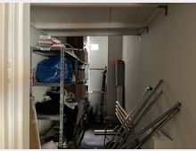 Ein kleiner Abstellraum mit weiß gestrichenen Wänden und einer niedrigen Decke. Links stehen Metallregale voller Gegenstände, darunter eine blaue Tasche, Kartons und Kleidung, rechts an der Wand angelehnt befinden sich Metallstangen. Im Hintergrund fällt durch ein schmales hohes Fenster etwas Tageslicht ein.