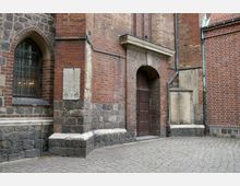 Backsteinfassade eines historischen Gebäudes mit einem rundbogigen Holzeingang, einem vergitterten Fenster und teilweise grobem Steinsockel. Der Boden vor dem Gebäude ist mit Pflastersteinen ausgelegt.