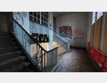 Verlassener Treppenaufgang in einem Gebäude mit Metallgeländern und großflächigen Graffiti an den Wänden; der Boden ist abgenutzt, und ein Teil der Fenster ist mit Holzplatten verbarrikadiert.